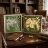 Une photographie réaliste et élégante, au format carré, présentant deux cadres lumineux en papier découpé sur le thème de la chasse, posés en évidence sur un bureau en bois verni de style classique. Les deux cadres partagent le même design dans les tons vert et blanc, représentant la silhouette d'un chasseur avec son fusil, un chien de chasse au premier plan, un cerf à l'arrière-plan, ainsi que des oiseaux en vol et de nombreuses petites étoiles. Le cadre de gauche montre le motif de jour avec un fond blanc éteint, tandis que le cadre de droite est illuminé de l'intérieur par une lumière jaune chaude et tamisée. Sur le bureau en cuir marron, on trouve également un carnet en cuir fermé, un stylo plume élégant, une boussole dorée, des livres anciens empilés, une carafe en cristal remplie de whisky avec son verre, et une plaque en laiton gravée. En arrière-plan, une bibliothèque en bois sombre remplie de livres et un fauteuil club en cuir marron complètent l'ambiance chaleureuse d'un bureau ou d'un salon d'adulte.