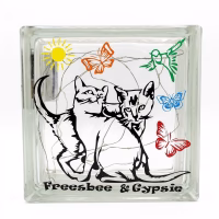 Bloc lumineux en brique de verre personnalisé pour chats. Le bloc affiche des silhouettes de deux chats, un soleil jaune, des papillons de différentes couleurs, un oiseau vert et le texte "Freesbee & Gypsie" gravé en bas. L'intérieur est rempli de guirlandes lumineuses à LED. Arrière-plan blanc uni