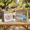 sac en toile de jute personnalisé. Deux sacs cabas en toile de jute personnalisés posés sur une table de jardin en bois. Le sac de gauche présente une impression florale avec une grande pivoine rose, et celui de droite une scène de plage avec mer bleue, étoile de mer et lunettes de soleil. En arrière-plan, un jardin fleuri avec des fleurs violettes et roses. cadoriginaux