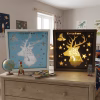 Une photographie réaliste et détaillée de deux cadres lumineux personnalisés en papier découpé, posés sur une commode en bois clair dans une chambre d'enfant. Le cadre de gauche, dans un encadrement en bois clair, montre un motif bleu avec un cerf blanc et des étoiles. Le cadre de droite, allumé dans un encadrement plus sombre, révèle le même motif de cerf et d'étoiles avec une lumière jaune dorée et chaleureuse. Le nom "Tristan" est découpé et illuminé au sommet des deux cadres. La commode est décorée avec un globe terrestre, une fusée jouet rouge, une pile de bandes dessinées et un petit robot rouge. En arrière-plan, on aperçoit une fenêtre avec des rideaux à motifs, un mur décoré d'affiches sur le thème de l'espace et des dinosaures, et un lit avec une couette à motifs de dinosaures. L'éclairage est doux et naturel, créant une ambiance chaleureuse et personnelle.