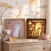 Une vue de face parfaitement cadrée, en format carré, d'une étagère de chambre d'enfant bien organisée. L'étagère en bois peint en blanc présente des objets de décoration assortis. Sur l'étagère du haut, on trouve de gauche à droite : un lapin en peluche blanc avec un nœud rose ; un ensemble de cinq blocs de bois naturels épelant 'L O V E' avec des cœurs ; une plante succulente en pot terracotta ; et une pile de livres d'enfants avec des titres clairs et lisibles en français ('Traces', 'Home', 'Le Petit Prince', 'Ki-Ki's'). L'étagère du bas contient, centrés côte à côte, deux cadres lumineux carrés artisanaux. Le cadre de gauche a un encadrement en bois clair et une découpe de papier 'Lina' avec un cerf gris et des étoiles. Le cadre de droite a un encadrement en bois sombre et la même découpe 'Lina' allumée, avec un cerf et des étoiles lumineux de couleur dorée et des étoiles chaudes. Le fond de l'étagère est un mur avec un motif doré de lune et d'étoiles. Le tapis floral détaillé est visible au premier plan, au bas de l'image. Le focus est net sur tous les objets, y compris tous les titres de livres et les textes de cadres, sous une lumière douce et naturelle.