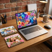 Tapis de souris personnalisable. Une photographie de style bureau à domicile montrant un ordinateur portable Dell argenté ouvert sur une surface en bois rustique. L'écran de l'ordinateur portable affiche un fond d'écran coloré d'art abstrait floral et aquarelle. Sur la gauche, deux tapis de souris rectangulaires sont empilés de manière oblique. Le tapis du dessus est bleu et présente une photo d'un homme âgé serrant dans ses bras deux enfants, avec le texte français « Bonne Fête Papi ! On t'aime ! Lucas & Jade ». Le tapis du dessous est rose et présente une photo d'une femme embrassant une jeune fille, avec le texte français « Bonne Fête Maman ! On t'aime ! Emma & Léa ». À droite, on voit une souris sans fil argentée et une tasse à café en céramique sur un sous-verre assorti au tapis de souris Maman. L'arrière-plan présente un mur en briques rouges apparentes, un porte-stylos rempli et une lampe de bureau. Le site Web « CadOriginaux.fr » est visible en bas à droite de l'écran de l'ordinateur portable.