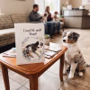 Protège carnet de santé animaux fait main. Dans une salle d'attente de clinique vétérinaire, un carnet de santé personnalisé est posé debout sur une table basse en bois. Il est blanc avec une couverture en cuir et porte l'inscription 'Carnet de santé Nougat' au-dessus d'une couronne de fleurs et d'empreintes de pattes, encadrant une photo d'un chiot Berger Australien bleu merle. Assis sur le carrelage à côté de la table, le vrai chiot Berger Australien regarde le carnet. En arrière-plan flou, un couple assis sur un canapé et un comptoir d'accueil sont visibles.