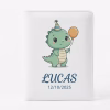 Protège-carnet de santé en cuir blanc, présenté en gros plan sur un fond blanc uni. La couverture est ornée d'une illustration de style dessin animé représentant un petit dinosaure vert portant un chapeau de fête rayé et tenant un ballon orange. En dessous de l'illustration, le prénom 'LUCAS' est écrit en lettres capitales bleu foncé, suivi de la date '12/10/2025'. Les bords du carnet sont cousus.