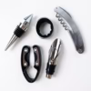 Gros plan des 5 accessoires de sommelier en métal argenté et détails noirs : un limonadier, un bouchon hermétique, un stop-goutte, un collier de bouteille et un découpe-capsule.