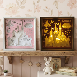 Une photographie d'ambiance montrant deux versions côte à côte d'une boîte à lumière en papier découpé personnalisée reposant sur une étagère en bois rustique dans une chambre d'enfant. Le fond est un papier peint à motif floral délicat de couleur rose pastel. La boîte de gauche possède un cadre en bois blanchi. À l'intérieur, les couches de papier découpé forment un paysage féerique blanc sur un fond rose : un château détaillé, une silhouette de fée, un croissant de lune, des nuages, des étoiles, des papillons, des oiseaux et des lapins. Le prénom « Esmée » et la date « 26.11.2025 » sont écrits en haut à droite. Cette version n'est pas allumée. La boîte de droite possède un cadre en bois brun foncé. Elle contient exactement le même design découpé, mais elle est allumée de l'intérieur par une lumière chaude et jaune doré. La lumière fait briller intensément les silhouettes et le nom, créant une ambiance magique et feutrée. Sous l'étagère, sur une surface blanche, on voit un lapin en peluche tricoté blanc, un livre de contes intitulé « Little Real Story Story » et une pile d'autres livres anciens. Une guirlande d'étoiles en bois est suspendue au bord de l'étagère.