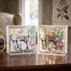Décoration luminaire personnalisé. Deux blocs de verre décoratifs posés sur une table en bois. Chaque bloc est illustré d'un dessin de deux chats en noir et blanc, entourés de papillons colorés, d'un oiseau et d'un soleil, avec l'inscription « Freesbee & Gypsie » en bas. Le bloc de droite est illuminé de l'intérieur par une guirlande lumineuse LED à micro-lucioles, tandis que celui de gauche est éteint.